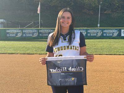 a photograph of Jamie Kelly posing with the catamounts climb higher banner, on the potomac state college softball field, in front of a background consisting of a fence and trim grass.  She is smiling and looking at the camera